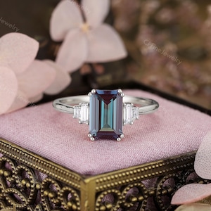 Puede incluir: Un anillo plateado con una gran gema rectangular que cambia de color. La gema cambia de verde azulado a p&uacute;rpura. Dos gemas blancas rectangulares m&aacute;s peque&ntilde;as flanquean la piedra principal. El anillo est&aacute; sobre una superficie rosa.