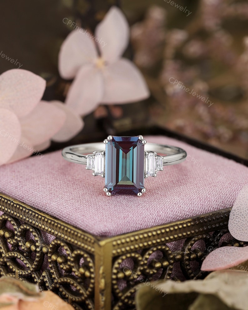 Puede incluir: Un anillo plateado con una gran gema rectangular que cambia de color. La gema cambia de verde azulado a p&uacute;rpura. Dos gemas blancas rectangulares m&aacute;s peque&ntilde;as flanquean la piedra principal. El anillo est&aacute; sobre una superficie rosa.