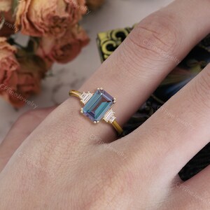 Puede incluir: Un anillo de oro con una piedra preciosa rectangular de color verde azulado en el centro, flanqueada por dos piedras blancas rectangulares m&aacute;s peque&ntilde;as. El anillo est&aacute; engastado en una delicada banda de oro, ofreciendo una est&eacute;tica cl&aacute;sica y elegante. El anillo est&aacute; en el dedo de una persona.