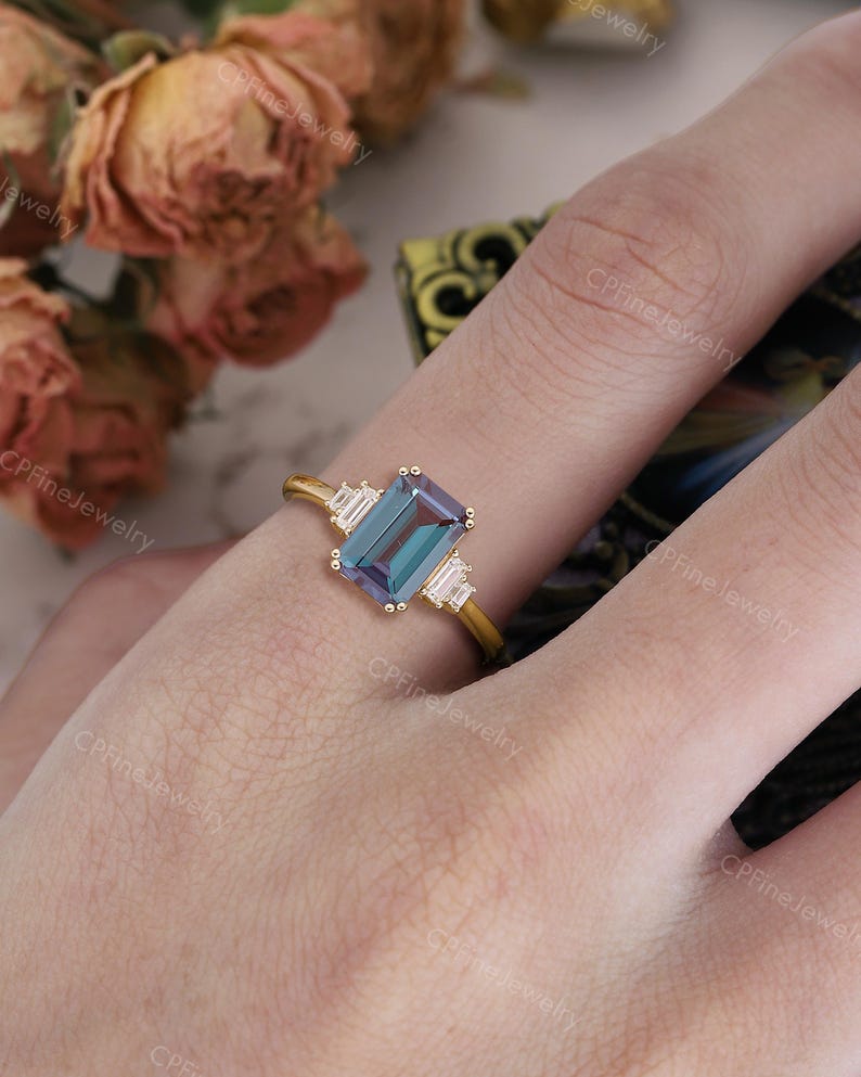Puede incluir: Un anillo de oro con una piedra preciosa rectangular de color verde azulado en el centro, flanqueada por dos piedras blancas rectangulares m&aacute;s peque&ntilde;as. El anillo est&aacute; engastado en una delicada banda de oro, ofreciendo una est&eacute;tica cl&aacute;sica y elegante. El anillo est&aacute; en el dedo de una persona.