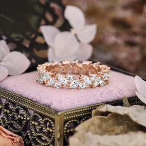 May include: Rose gold eternity ring featuring marquise-cut white gemstones. The ring is set on a pink surface inside a decorative box. The background includes flowers and the text "CPFineJewelry".