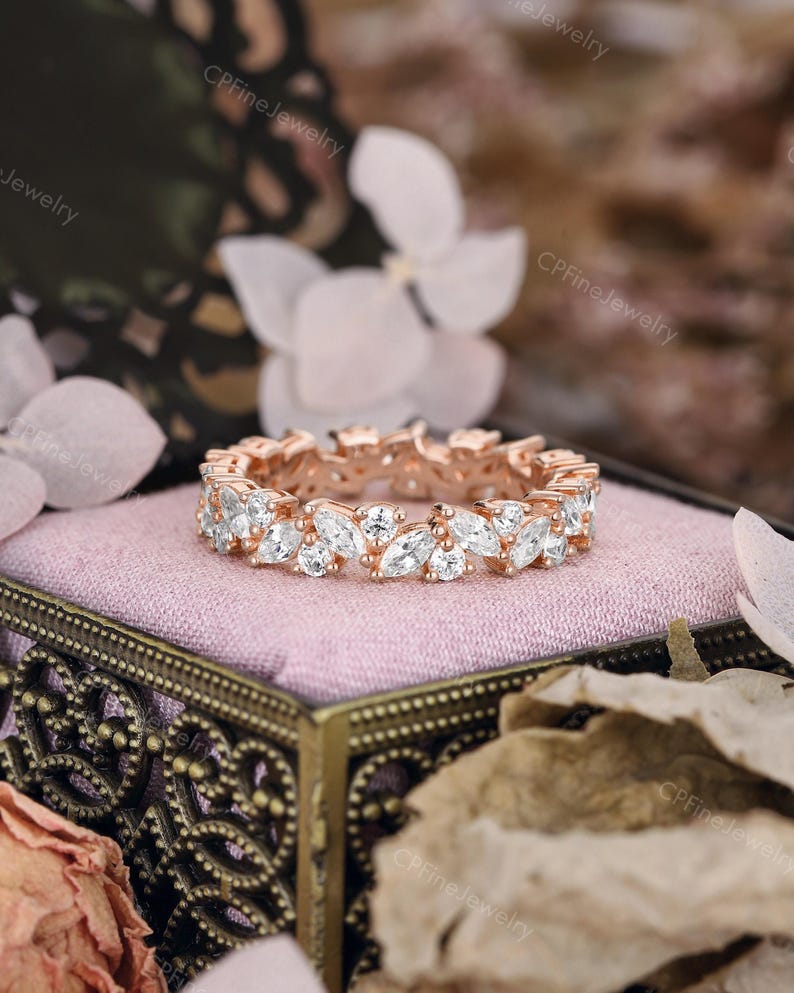 May include: Rose gold eternity ring featuring marquise-cut white gemstones. The ring is set on a pink surface inside a decorative box. The background includes flowers and the text "CPFineJewelry".