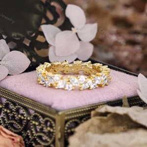 May include: A gold-colored ring with a continuous pattern of clear, marquise-cut gemstones. The ring is set in a gold band and rests on a pink velvet surface inside a decorative box. The background includes white flowers and foliage.