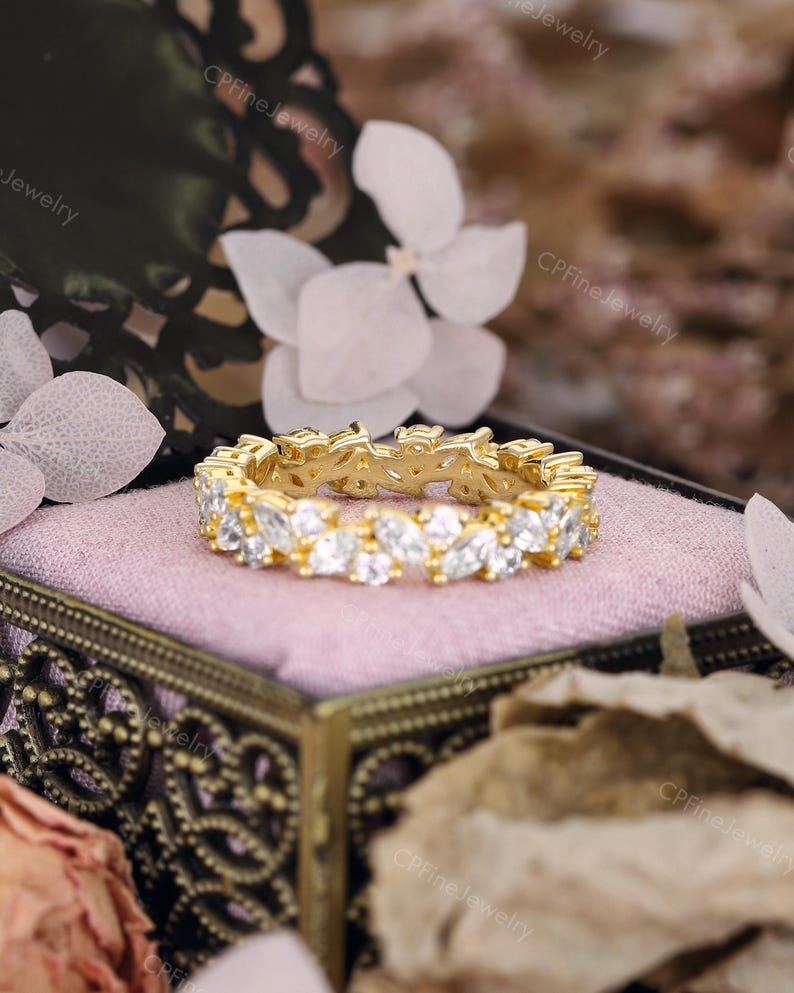 May include: A gold-colored ring with a continuous pattern of clear, marquise-cut gemstones. The ring is set in a gold band and rests on a pink velvet surface inside a decorative box. The background includes white flowers and foliage.