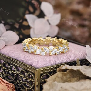 May include: A gold-toned ring with a continuous pattern of marquise-cut white gemstones. The ring is set on a pink surface inside a decorative box. The background features soft focus flowers and foliage, creating a romantic aesthetic. The ring is a piece of jewelry.