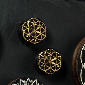 May include: Two black wooden round ornaments with gold geometric designs. The designs are intricate and detailed, and the gold paint is shiny. The ornaments are on a black background.