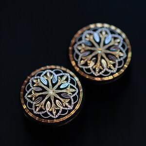 May include: Two round, black, decorative buttons with gold and silver filigree designs. The buttons have a floral pattern.