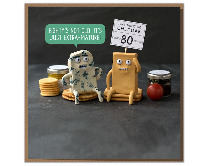 K&ouml;nnte beinhalten: Zwei Cartoon-K&auml;sefiguren, ein Blauschimmelk&auml;se und ein Cheddar, sitzen auf Crackern mit einer Sprechblase &uuml;ber dem Blauschimmelk&auml;se, die sagt "Eighty's not old. It's just extra-mature!" und einem Schild &uuml;ber dem Cheddar, das sagt "Fine Vintage Cheddar Aged 80 Years".