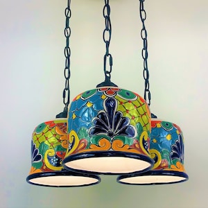 May include: A three-light chandelier with colorful, hand-painted ceramic shades. The shades feature a floral pattern in shades of blue, yellow, green, and red. The chandelier is suspended from a black metal ceiling plate with three chains.