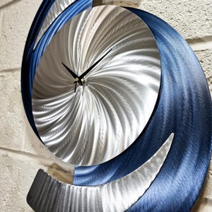May include: A modern wall clock with a silver and blue metal design. The clock face has a swirling pattern and black hands.