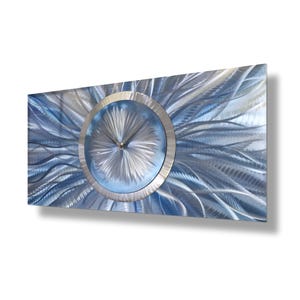 Może przedstawiać: Nowoczesny zegar ścienny o metalowym wykończeniu w kolorze niebieskim i srebrnym. Tarcza zegara jest otoczona srebrnym pierścieniem i ma srebrne wskazówki.