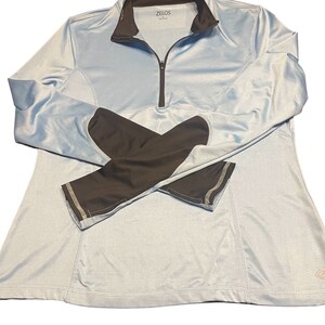 May include: Light blue athletic top with black sleeves and a quarter-zip closure. The top features the brand name "ZELOS" on the collar. The sleeves have a contrasting gray trim. The top is designed for athletic activities.
