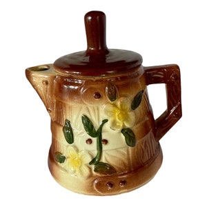May include: A brown and cream-colored ceramic teapot with a lid and handle. The teapot is decorated with embossed flowers and leaves. The handle and lid are a darker shade of brown. The teapot has a spout and is designed to hold liquid.