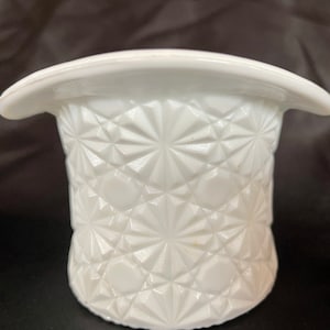 May include: A white, vintage-style top hat-shaped container. The container has a textured, starburst pattern on the sides and a smooth, curved brim. The object is made of opaque white glass or ceramic.