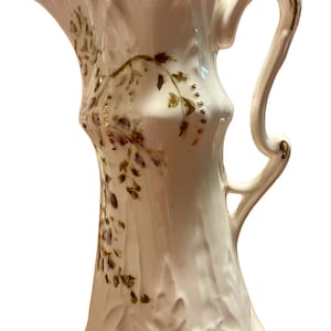 May include: A white ceramic pitcher with a ruffled rim and a curved handle. The pitcher is decorated with a floral design in shades of brown and gold accents. The rim and handle are trimmed with gold.