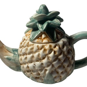 May include: A ceramic pineapple-shaped teapot. The body is cream-coloured with a textured surface, complemented by green leaves, handle, and spout. The lid, also green, mirrors the pineapple crown.