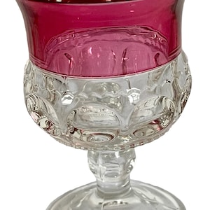 May include: A clear glass goblet with a ruby red rim. The goblet features a detailed, embossed design on the lower half of the bowl. The stem and base are also clear glass, creating a classic, elegant look.