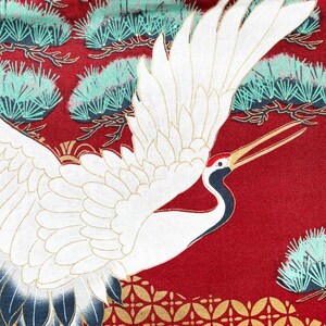 Puede incluir: Una gr&uacute;a blanca con las alas extendidas sobre un fondo rojo con un patr&oacute;n dorado y verde. La gr&uacute;a tiene un pico largo y delgado y una mancha roja en la cabeza.