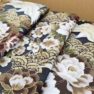 以下が含まれることがあります： 日本のデザインにインスパイアされた折り畳まれた生地のクローズアップ。生地は、白い花、鶴、金色のアクセントが黒い背景に特徴です。デザインは詳細で複雑で、豪華な雰囲気です。