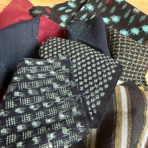 Può includere: Una collezione di ritagli di tessuto assortiti con vari motivi e texture. I pezzi includono tessuti blu scuro, neri e bordeaux con stampe geometriche, a griglia e in stile ikat nei toni del bianco, azzurro e marrone terroso.