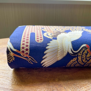 May include: Blue fabric with a white crane design. The crane is flying with its wings spread. The fabric has a gold and red pattern.