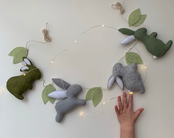 Puede incluir: Una guirnalda decorativa con conejitos de fieltro en tonos verdes y grises, intercalados con hojas verdes de fieltro. Los conejitos tienen detalles blancos y están ensartados en una guirnalda de luces. La mano de un niño se extiende hacia la guirnalda.