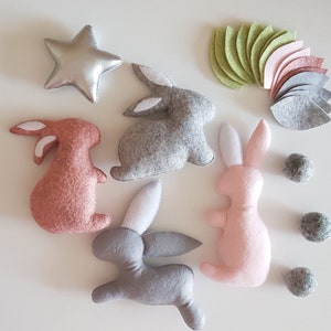 May include: A set of felt bunny toys in pink, gray, and brown with white accents. The bunnies are arranged on a white surface with felt flower petals and small felt balls.