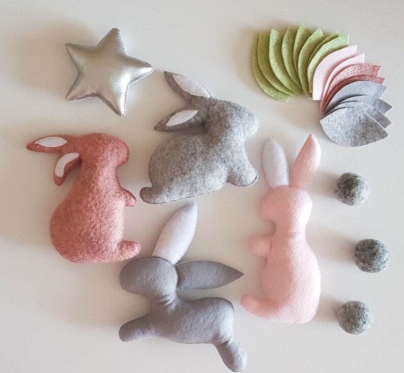 May include: A set of felt bunny toys in pink, gray, and brown with white accents. The bunnies are arranged on a white surface with felt flower petals and small felt balls.