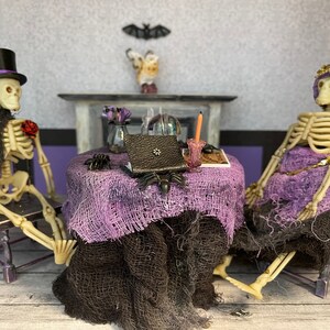 May include: A Halloween-themed diorama featuring two skeleton figures seated at a table. The skeletons are dressed in formal attire, with one wearing a top hat. The table is draped with purple fabric and adorned with a spider and a book. A candle and a small vase of flowers are also present.