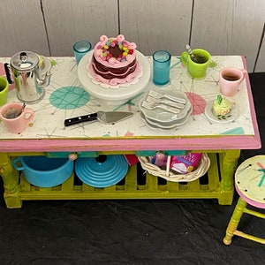 May include: A miniature kitchen scene with a pink and green table, a chocolate cake, and various dishes. The table has a patterned surface with a pink border. A small stool is next to the table. The scene includes a teapot, cups, and plates.