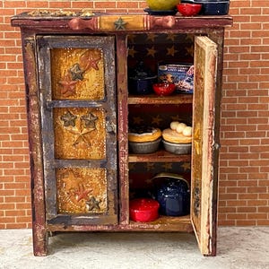 May include: A miniature, rustic-style cabinet with a distressed finish. The cabinet features a door with star cutouts and is filled with miniature kitchen items, including bowls, pies, and a cookbook. The cabinet is painted in shades of red, brown, and gold.