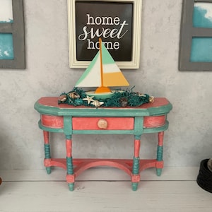 May include: A miniature coral and teal painted table with a drawer, adorned with a sailboat, seashells, and a decorative net. The table is set against a wall with a framed "home sweet home" sign and window art.
