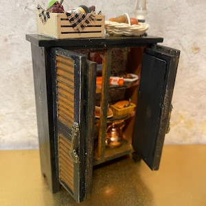 May include: Miniature black cabinet with open doors, revealing shelves stocked with food items. A wooden crate with grapes and a bottle of wine sits atop the cabinet, alongside a basket of eggs and a small lamp. The cabinet doors have a wooden slat design.