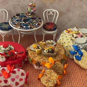 May include: Miniature dollhouse furniture and accessories. Includes a mosaic-tiled table with two white chairs, and several small tables with cakes, pastries, and tea sets. The tables are covered with patterned tablecloths, and the scene is set on a brick-patterned surface.