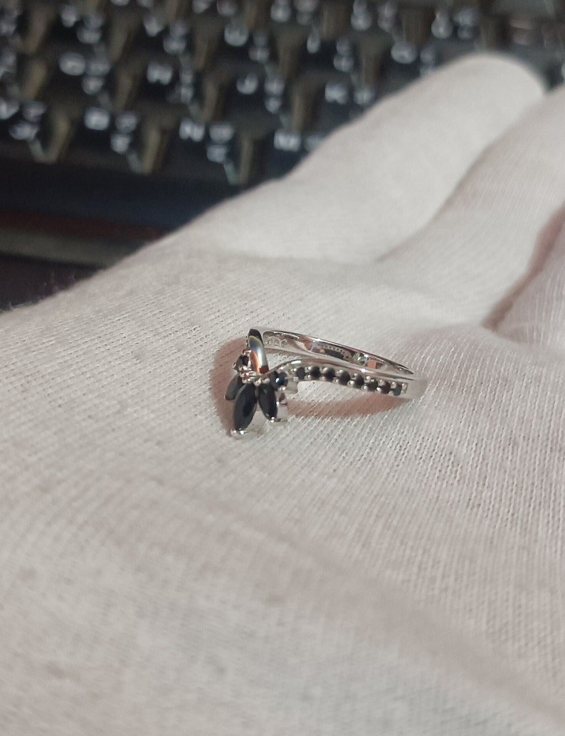 May include: A silver ring with a V-shaped design and black gemstones. The ring is set with small black stones along the band and larger black stones in the V-shaped design.