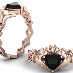 Puede incluir: Un anillo de nudo celta de oro rosa con una piedra preciosa negra en forma de corazón. El anillo presenta dos manos entrelazadas, simbolizando el amor y el compromiso.