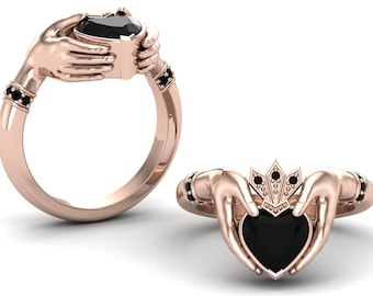 May include: Rose gold Claddagh ring featuring a heart-shaped black gemstone held by two hands. The ring has a crown above the heart and black gemstones on the band. The design is a symbol of love, loyalty, and friendship.