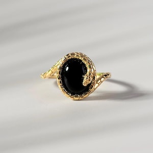 May include: Gold ring with an oval black stone, framed by a textured band. The ring's design includes a snake motif, with the snake's head and body encircling the stone. The ring is set against a white background.