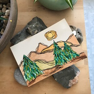 May include: Watercolour painting of a mountain landscape with three green trees and a yellow sun. The mountains are brown and the sky is white.