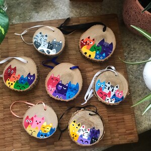 May include: Seven circular wood slice ornaments, each featuring a hand-painted cluster of colorful cats. The ornaments have natural bark edges and are attached to thin ribbons in various colors for hanging. Each piece displays a unique arrangement of feline faces in shades of red, orange, yellow, green, blue, purple, pink, and grey.