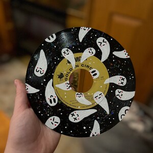 May include: A black vinyl record painted with white ghosts with pink cheeks and black eyes. The record has a black and white speckled background. The record label is gold with the text "GOLDEN CIRC" and "VIC" in black.