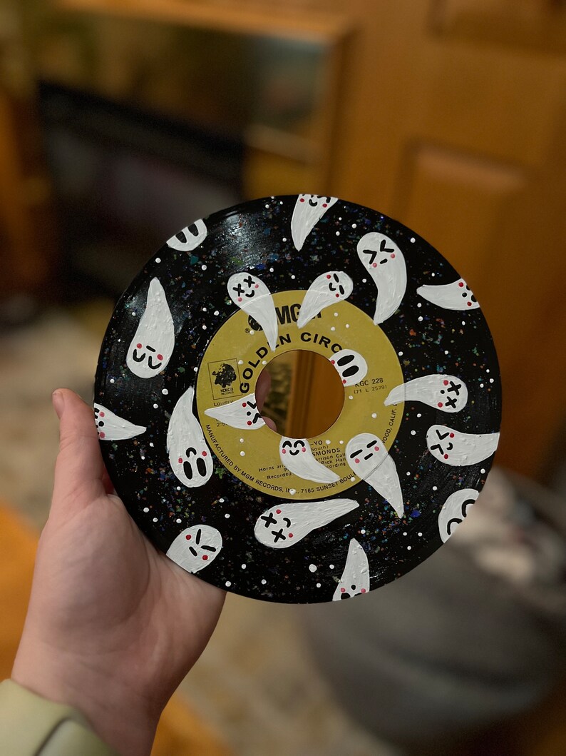 May include: A black vinyl record painted with white ghosts with pink cheeks and black eyes. The record has a black and white speckled background. The record label is gold with the text "GOLDEN CIRC" and "VIC" in black.