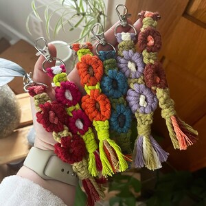 May include: A set of six colourful crocheted keychains with flower designs. Each keychain has a silver key ring and a tassel. The keychains are made with different coloured yarn, including red, orange, yellow, green, blue, and purple.