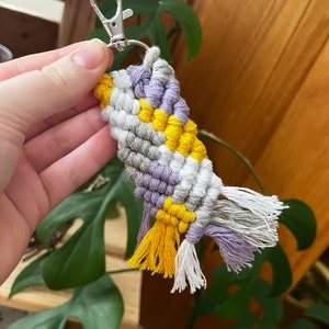 May include: A purple, white, and yellow macrame keychain with a silver key ring. The keychain has tassels on the bottom.