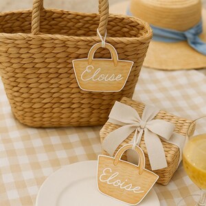 May include: A woven basket with rope handles, a straw hat with a blue ribbon, and a gift box with a bow sit on a checkered tablecloth. A paper cutout of a purse with the name "Eloise" is attached to the basket and gift box.