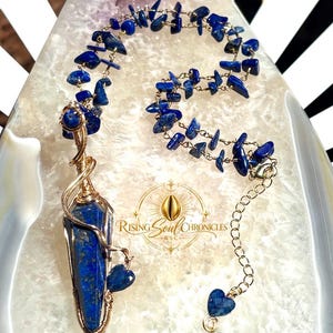 May include: A wire-wrapped necklace featuring a pointed lapis lazuli pendant and a chain made of small, irregular lapis lazuli chips. The metal wire is gold-toned. A small heart-shaped lapis lazuli charm hangs from the pendant and the chain end.