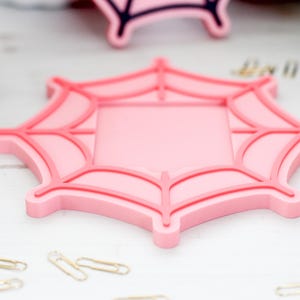 May include: A pink spiderweb-shaped coaster with a square center. The coaster has a raised, darker pink outline and web details. Several gold paperclips are scattered around the coaster on a white surface.