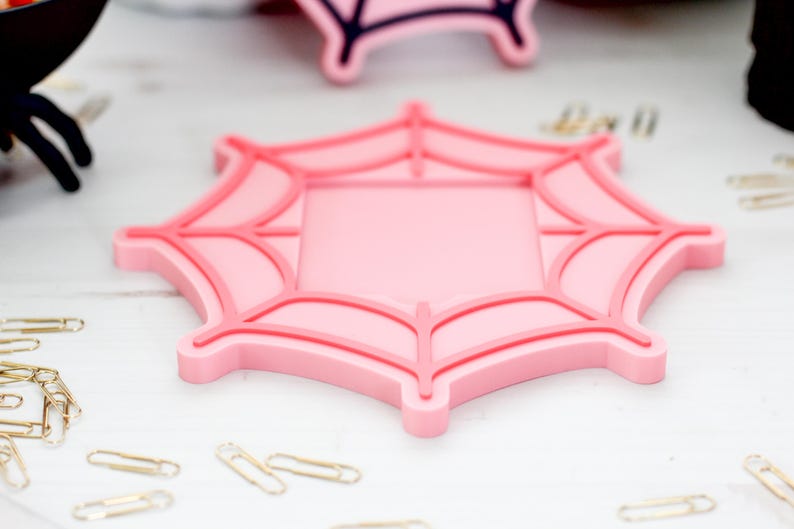 May include: A pink spiderweb-shaped coaster with a square center. The coaster has a raised, darker pink outline and web details. Several gold paperclips are scattered around the coaster on a white surface.