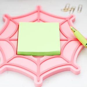 Op de afbeelding: Een roze spinnenwebvormige bureau-organizer bevat een stapel lichtgroene plaknotities. Een lichtgroene pen met gouden accenten ligt op de organizer. Er liggen verschillende gouden paperclips verspreid.