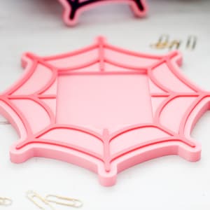 May include: A pink spiderweb-shaped coaster with a square center. The coaster has a raised, darker pink outline and web details. Gold paperclips are scattered around the coaster, suggesting a Halloween or spooky theme.